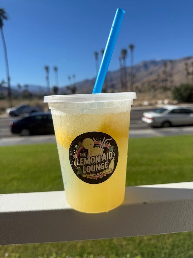 Plastic cup with yellow drink and blue straw on a railing, with a road and mountains in the background