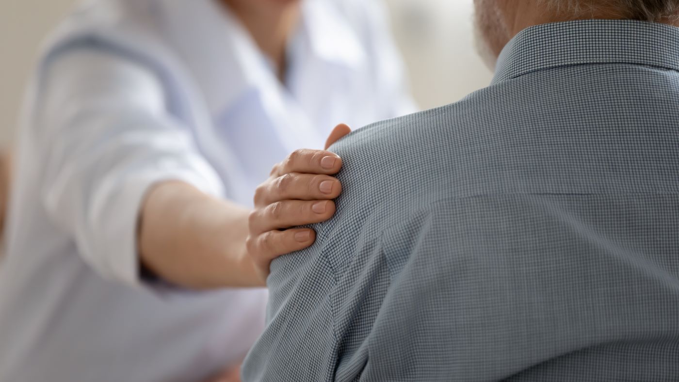 A medical professional in a white coat comforts a patient, hand on their shoulder.
