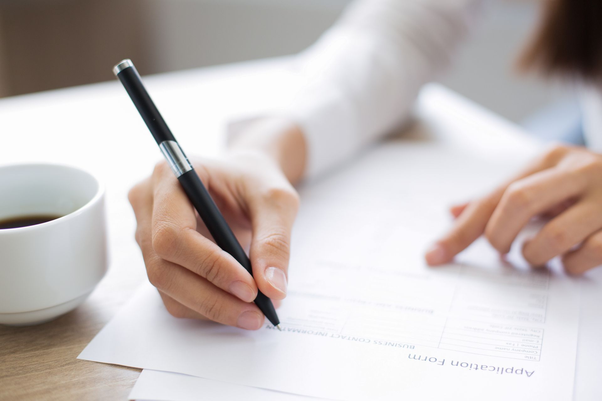 A person's hand holding a pen, writing on a form at a desk, next to a coffee cup.