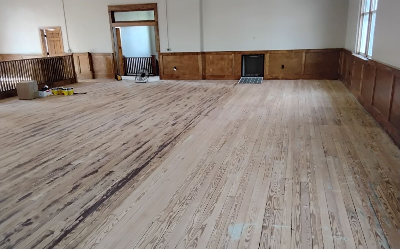 A light-wood floor being sanded in a large room with wood paneling on the walls and natural light from a window.