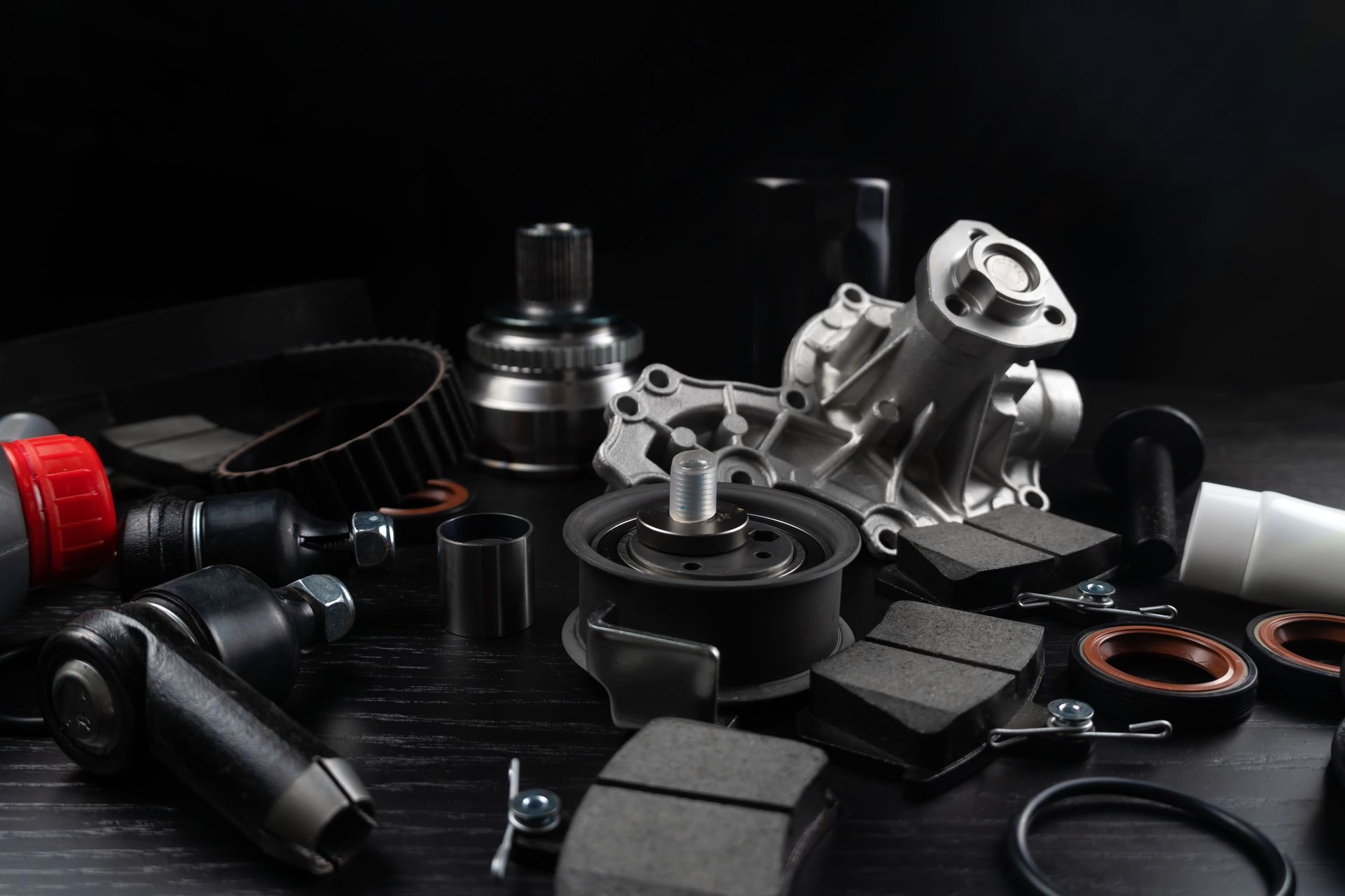 Parti di automobili su una superficie scura, tra cui una pompa dell'acqua, pastiglie dei freni e cinghia di trasmissione.