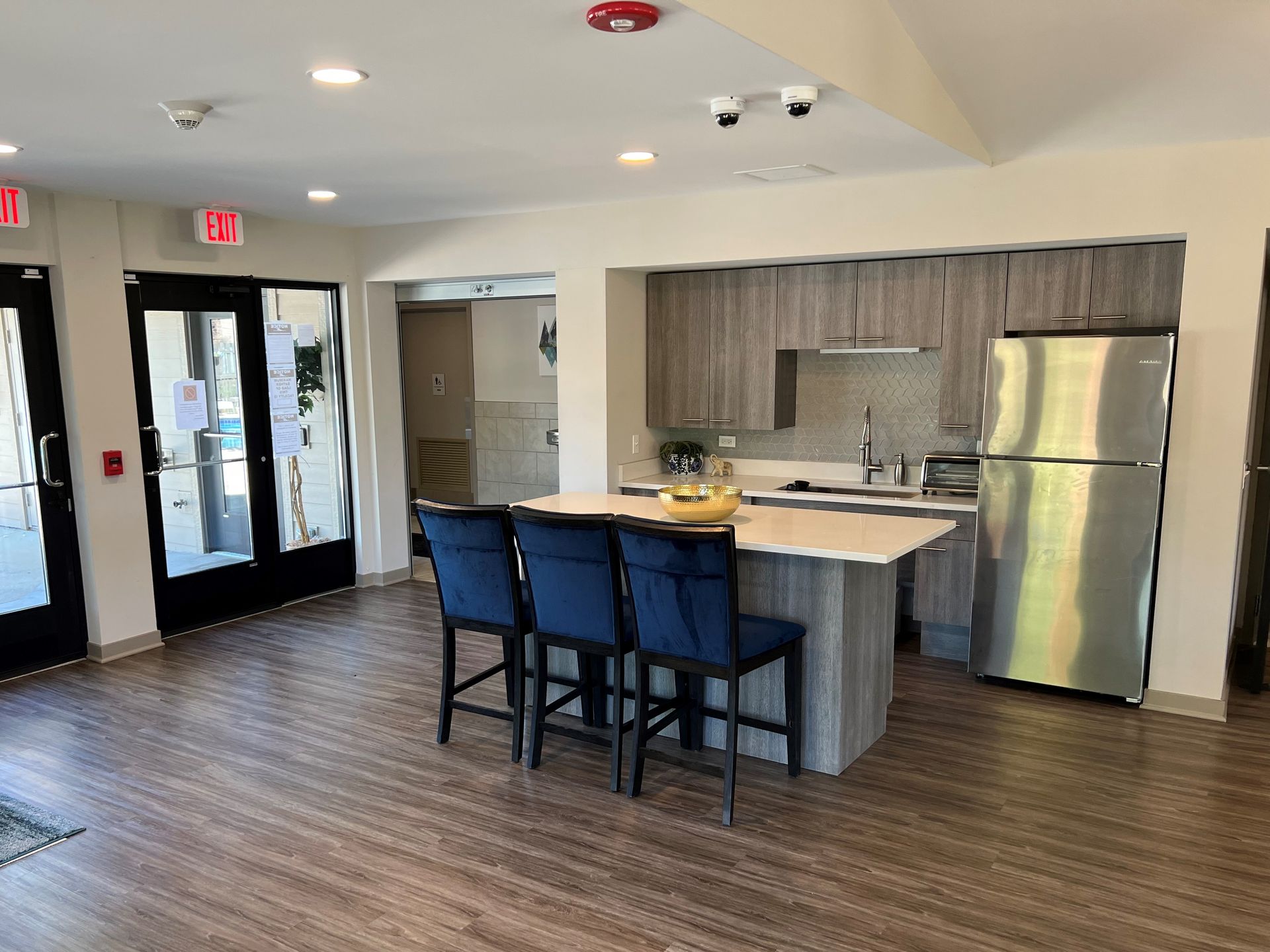 Kitchen area with a breakfast bar and blue chairs, next to stainless steel appliances and exit doors.