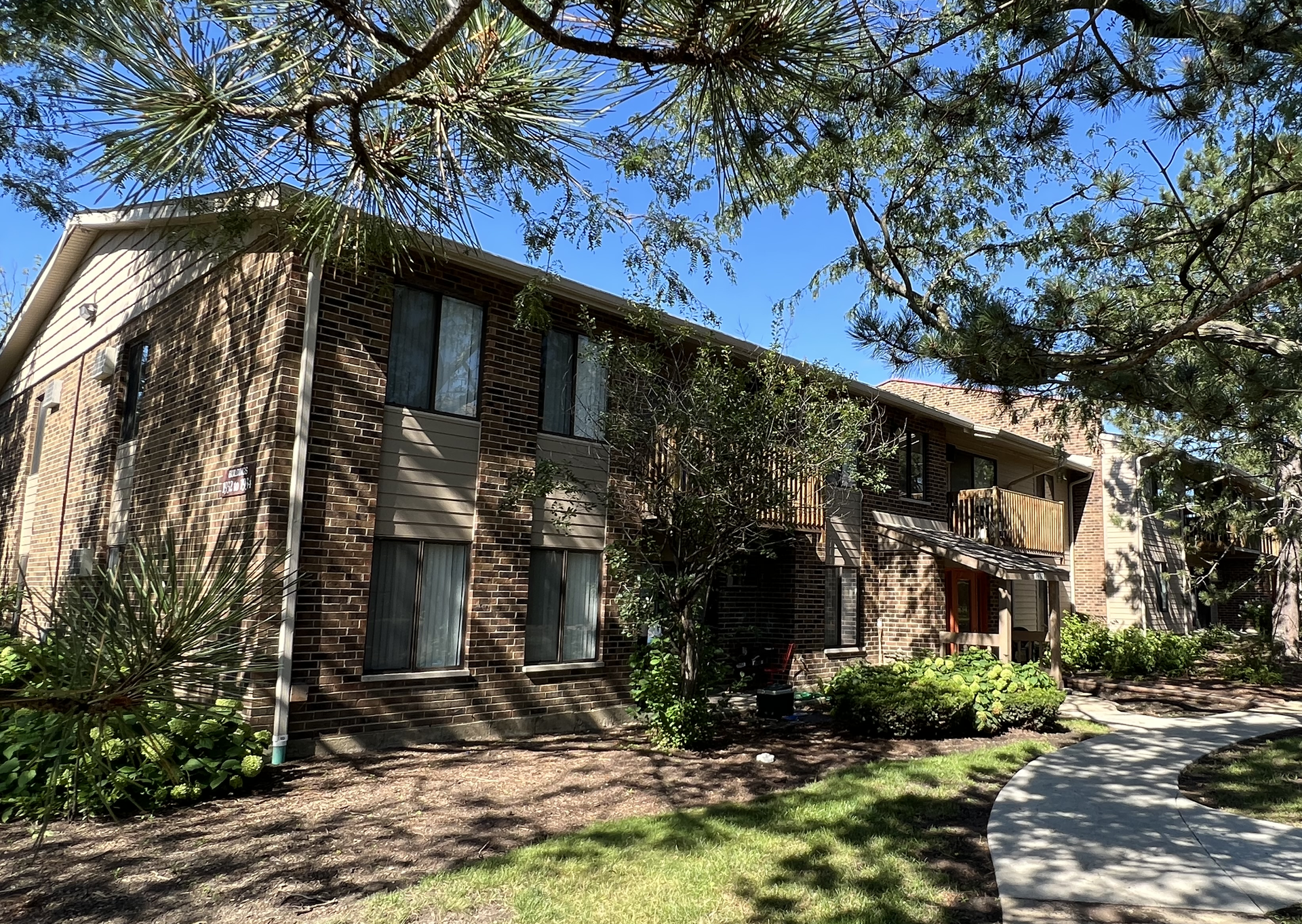 Two-story brick apartment building on a sunny day, framed by green trees and foliage.
