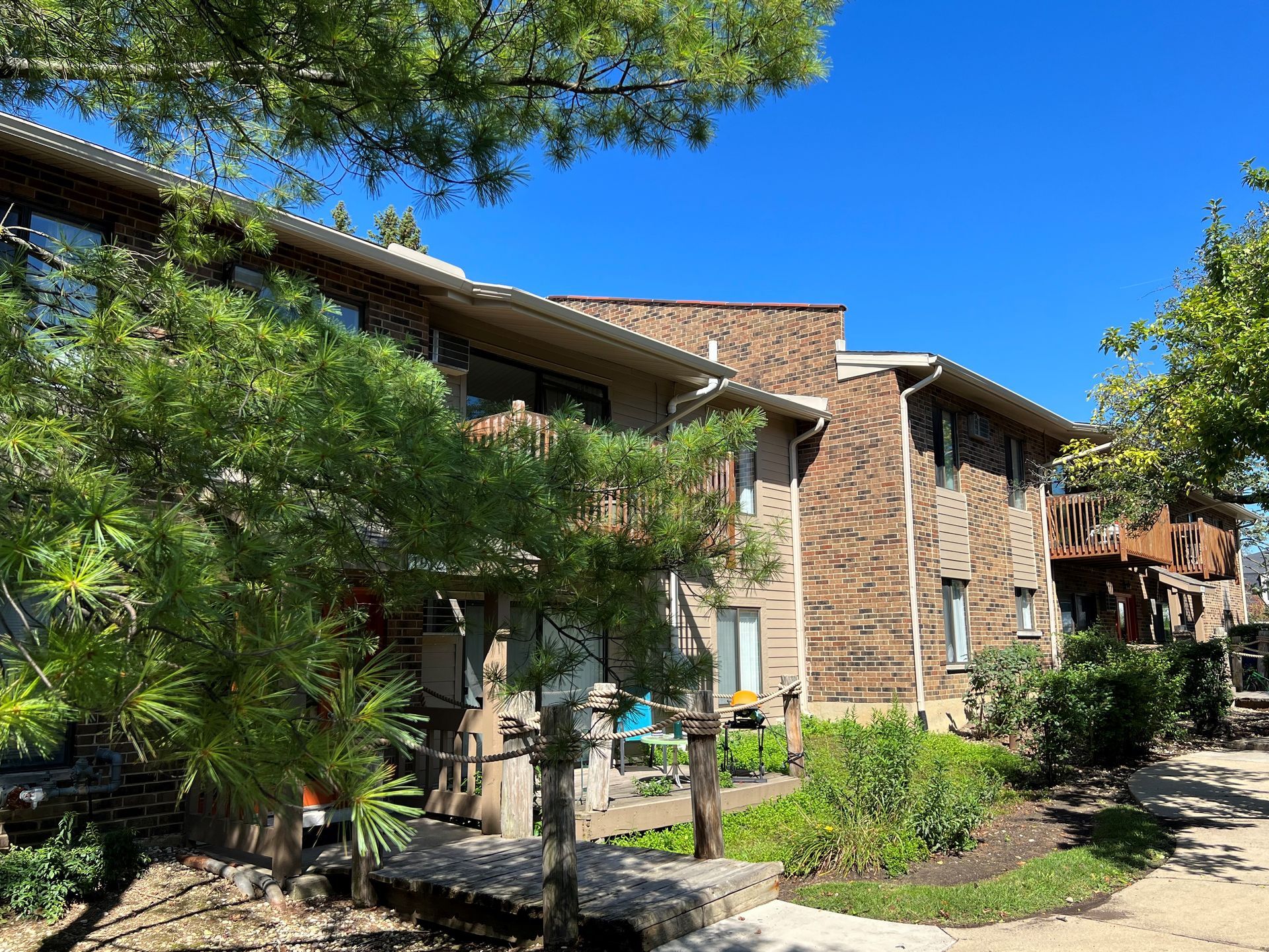 Two-story apartment buildings with brick and brown siding, trees in front, under a blue sky.