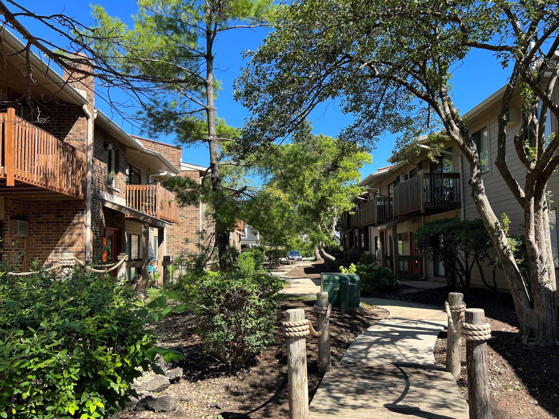 Apartment buildings with wooden balconies, pathway, and lush trees under a bright blue sky.