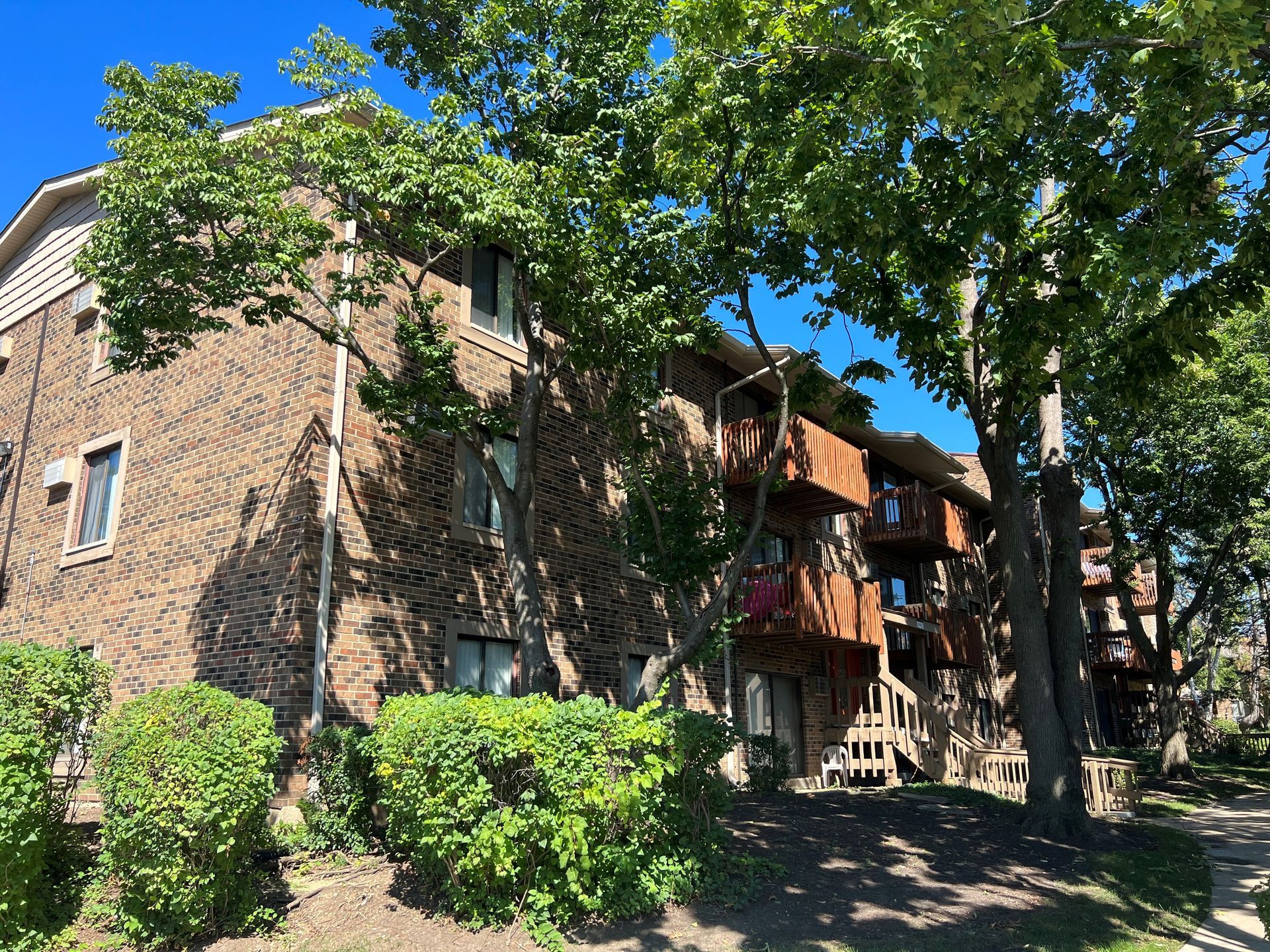 Brick apartment building with balconies, partly shaded by trees, sunny day.
