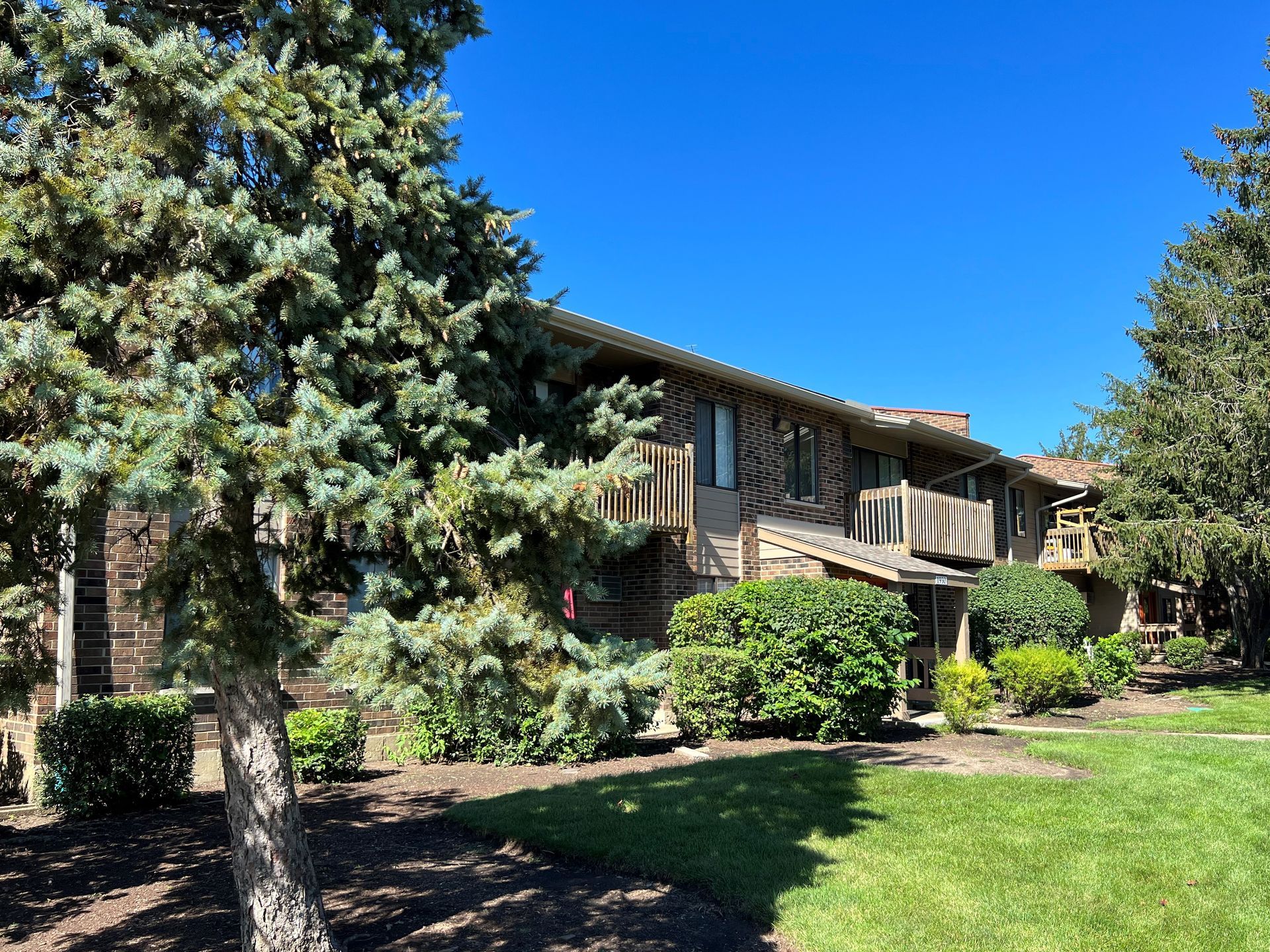 Two-story brick apartment building with balconies, surrounded by trees and green lawn under a blue sky.