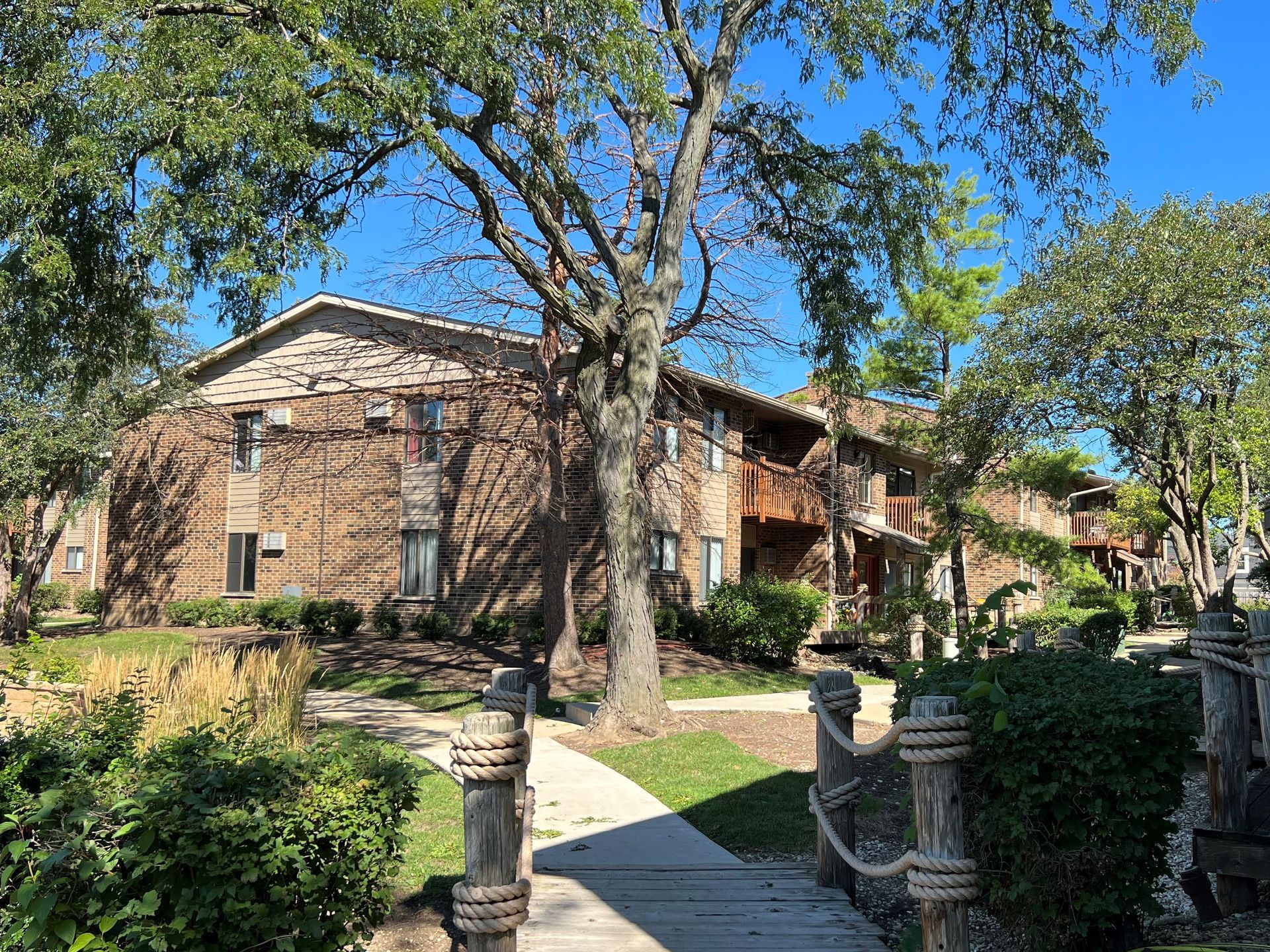 Apartment building with brick exterior, nestled among trees, overlooking a small bridge.