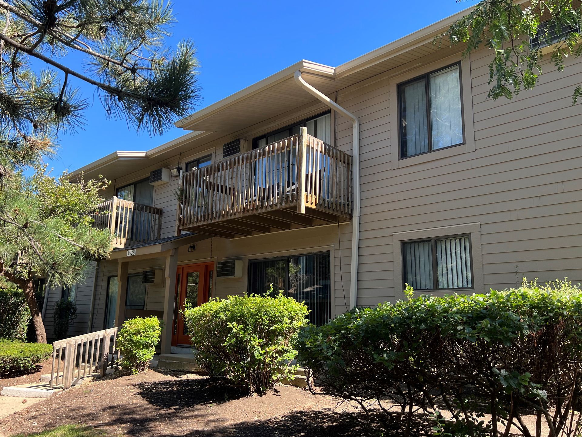 Two-story beige apartment building with balconies, brown railings, and shrubs in front.