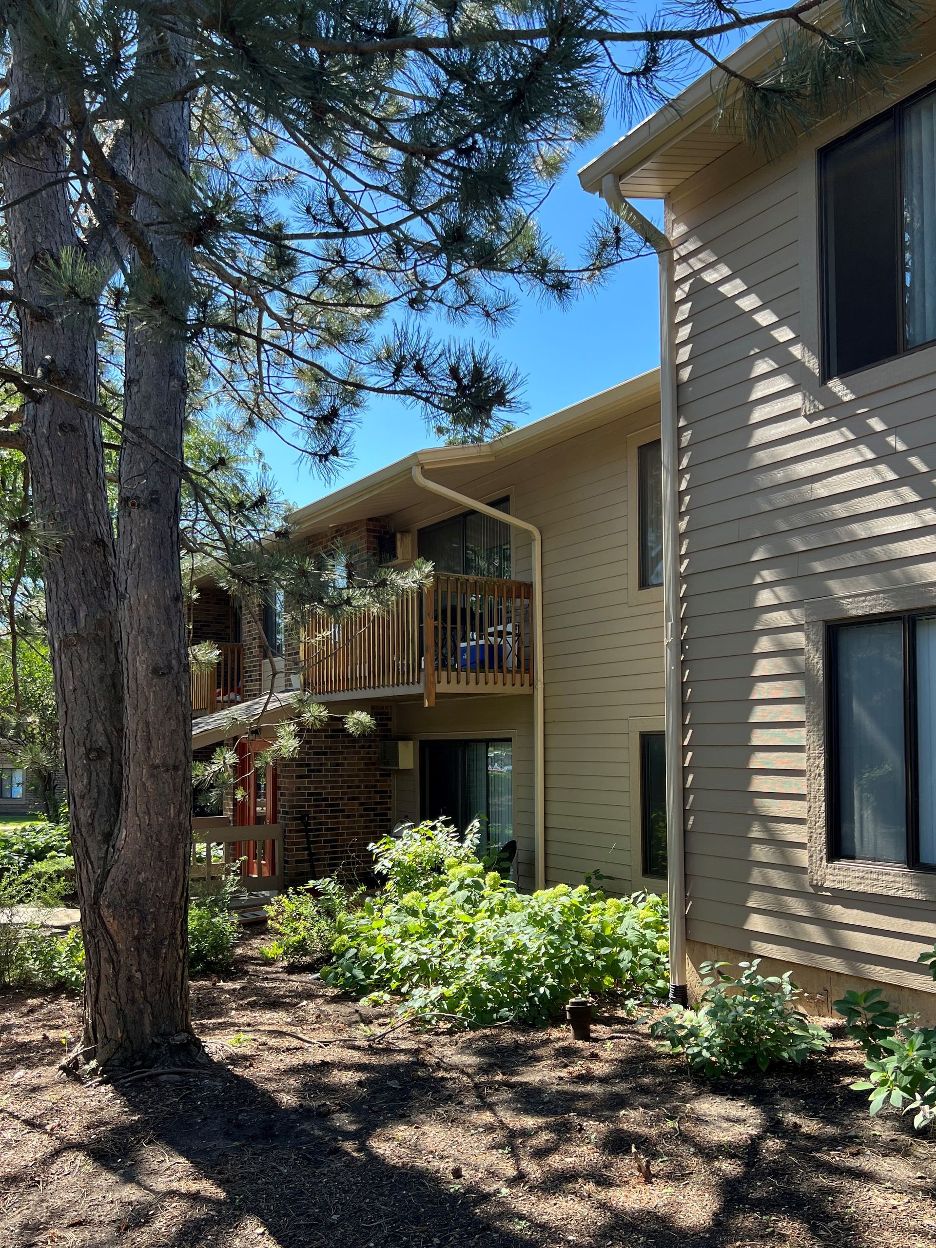 Beige apartment building with a wooden balcony surrounded by trees and greenery under a blue sky.
