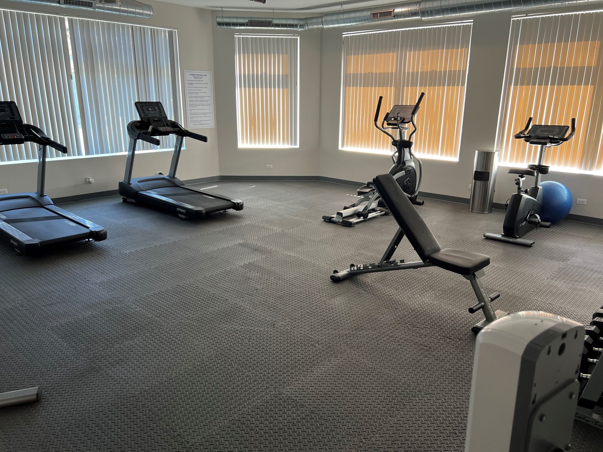 Gym with treadmills, exercise bikes, weight bench, and windows with blinds. Black flooring.