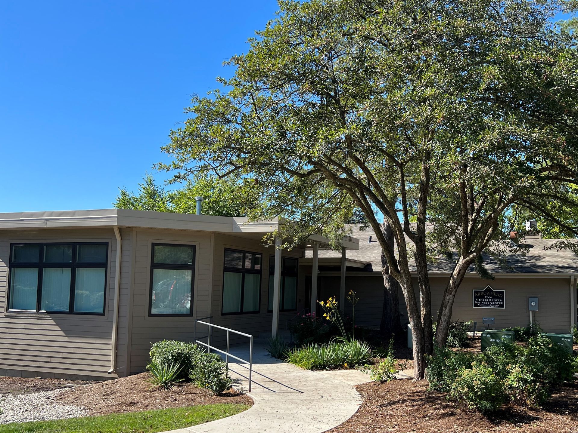 Exterior of a one-story building with a walkway, surrounded by greenery and trees; blue sky.
