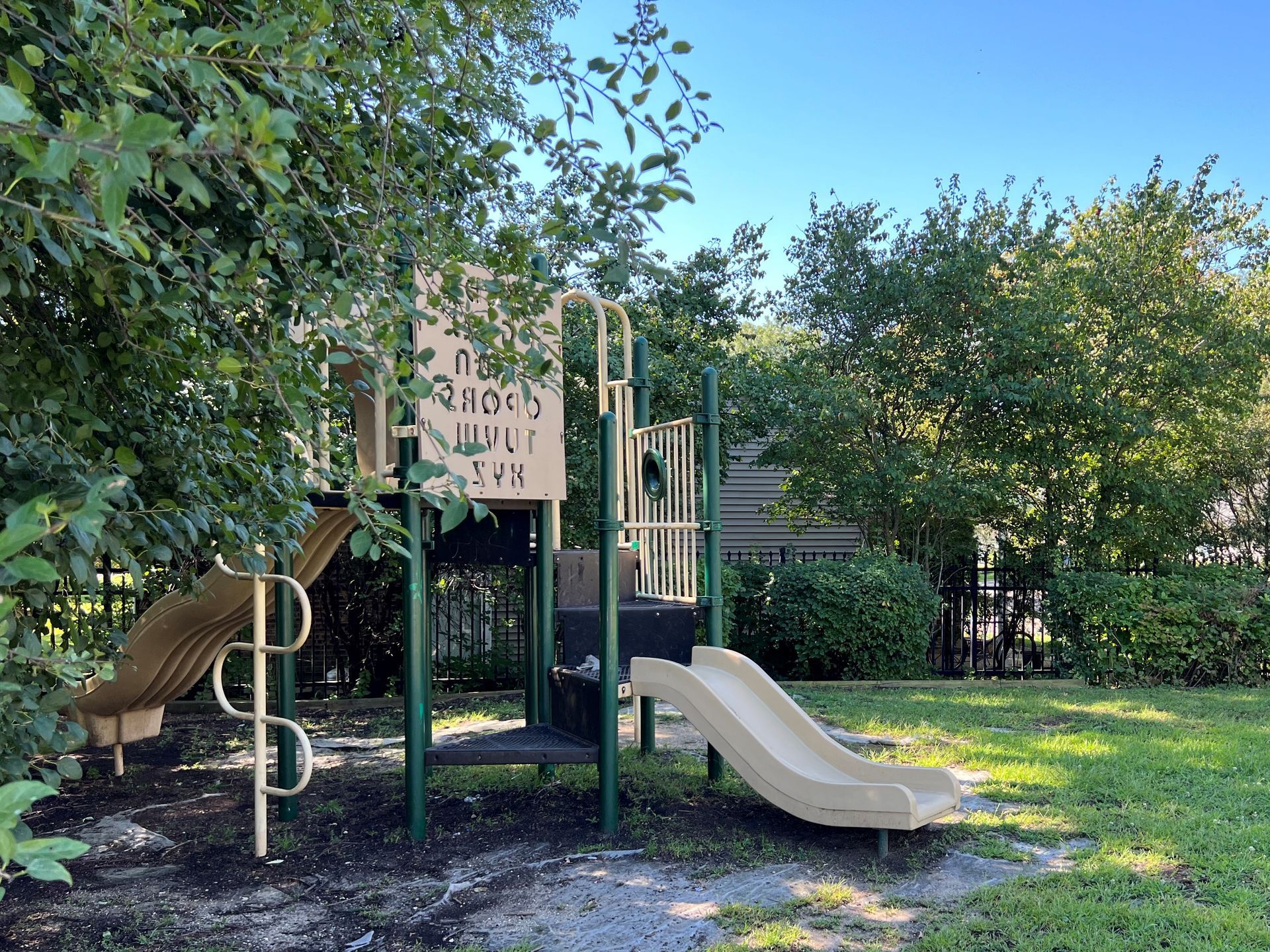 Playground with two slides, green support beams, and tan plastic. Surrounded by trees and grass.