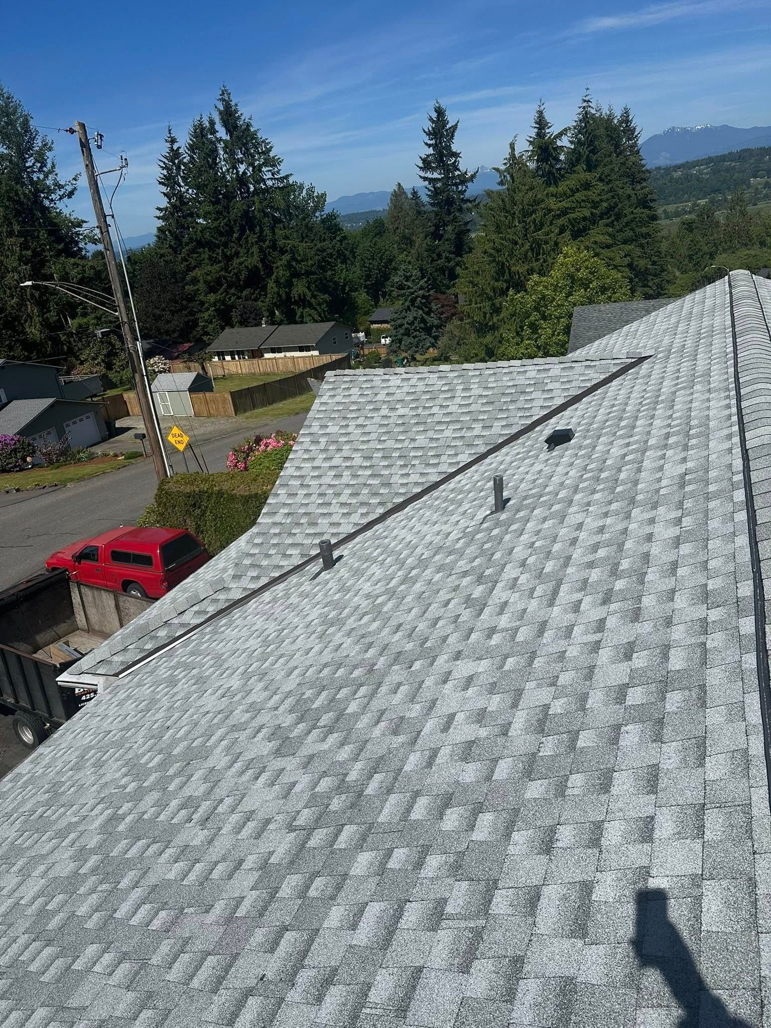 Gray shingle roof with a pipe, with trees and a red truck in the background on a sunny day.