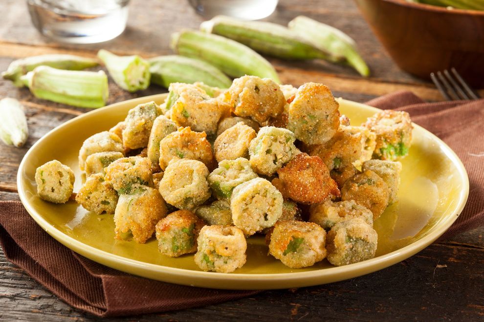 Fried okra on a yellow plate, with fresh okra in the background. The okra is golden-brown, with a wooden table setting.
