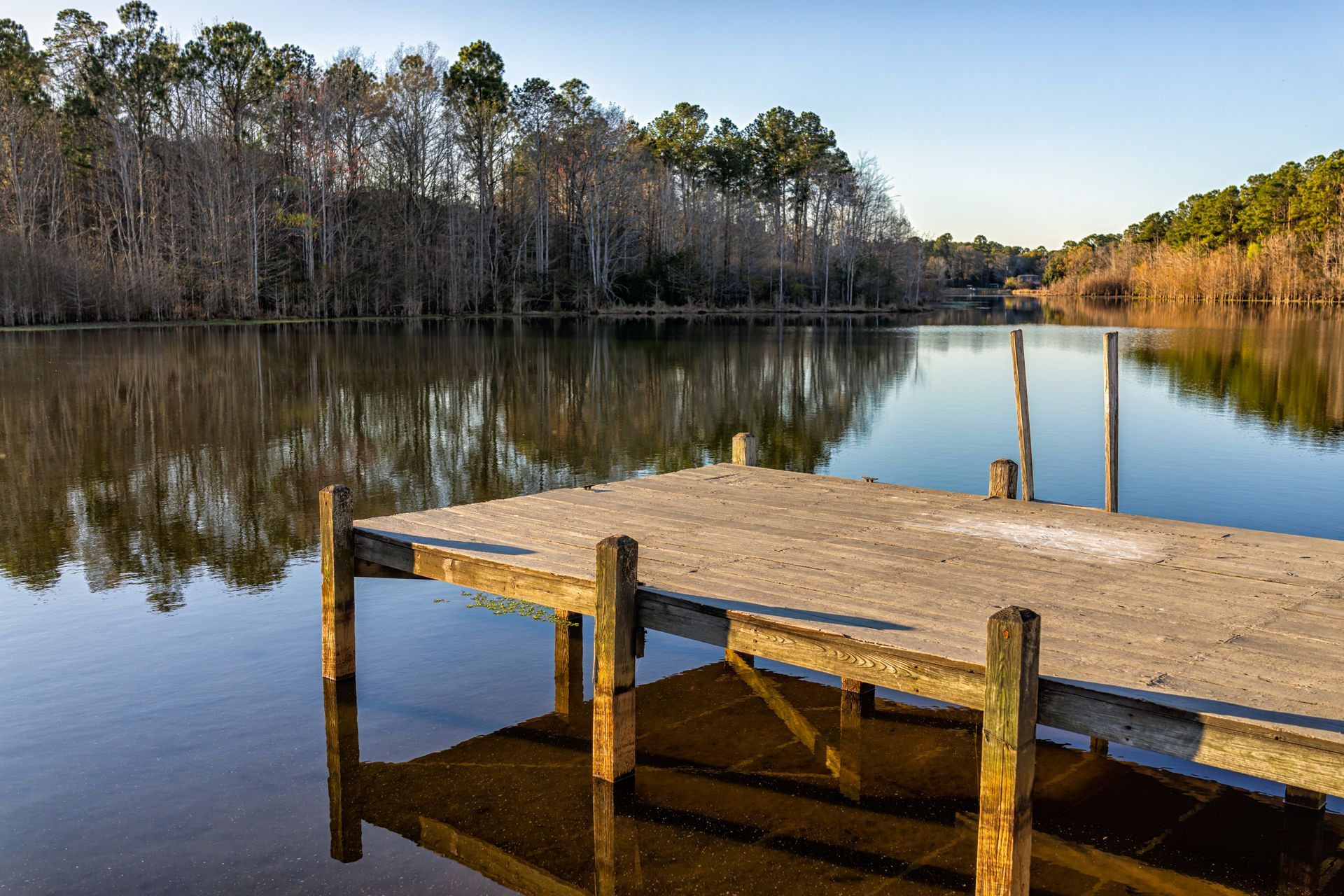 A weathered wooden dock extends into a calm lake with trees lining the shore under a clear sky.