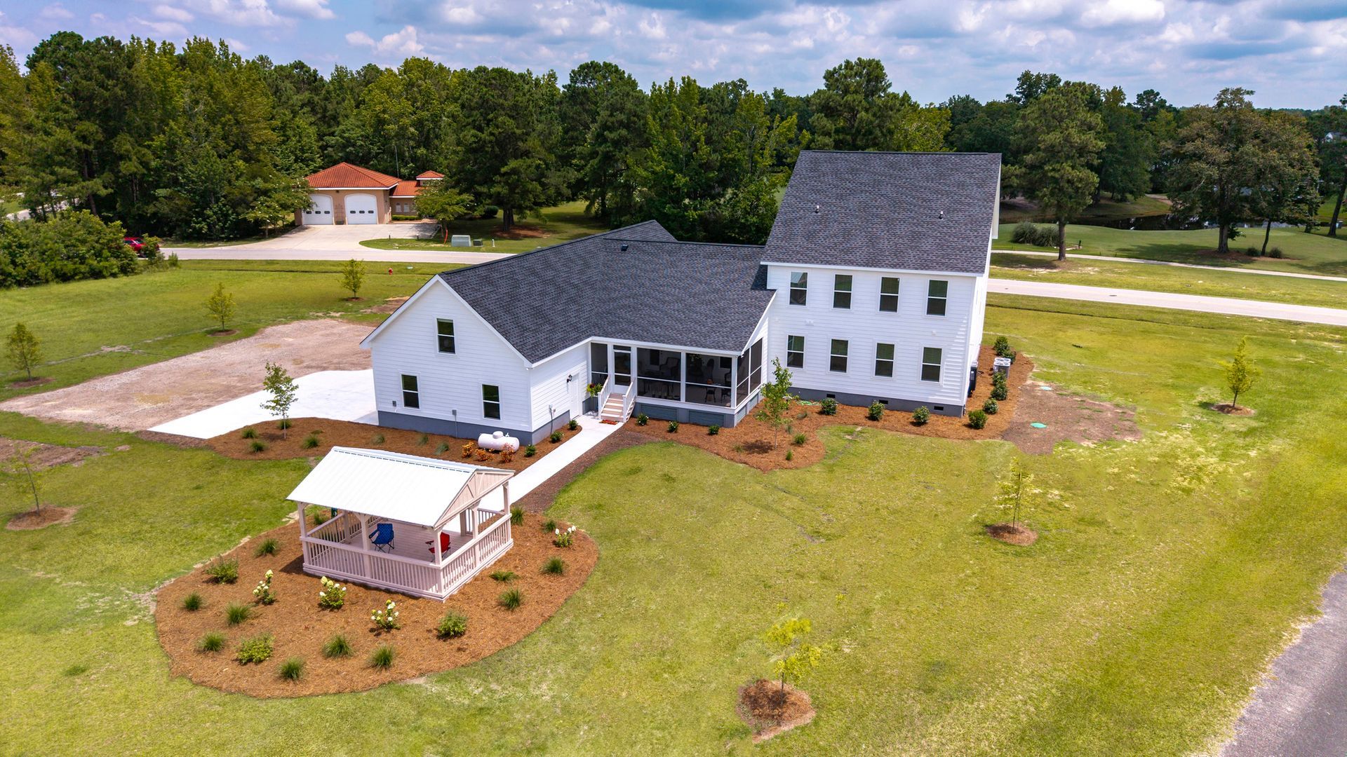 Aerial view of a white house with a black roof and a small playhouse. The house is surrounded by a lawn with trees in the background.