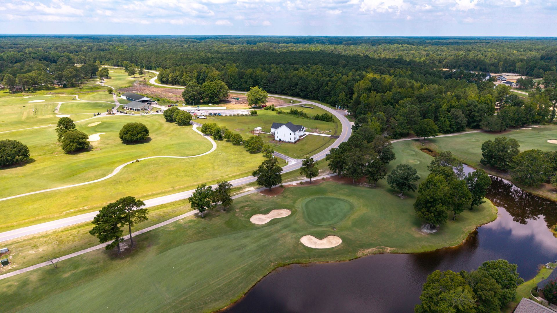 Aerial view of a golf course with green fairways, sand traps, and a lake surrounded by trees and a white house.