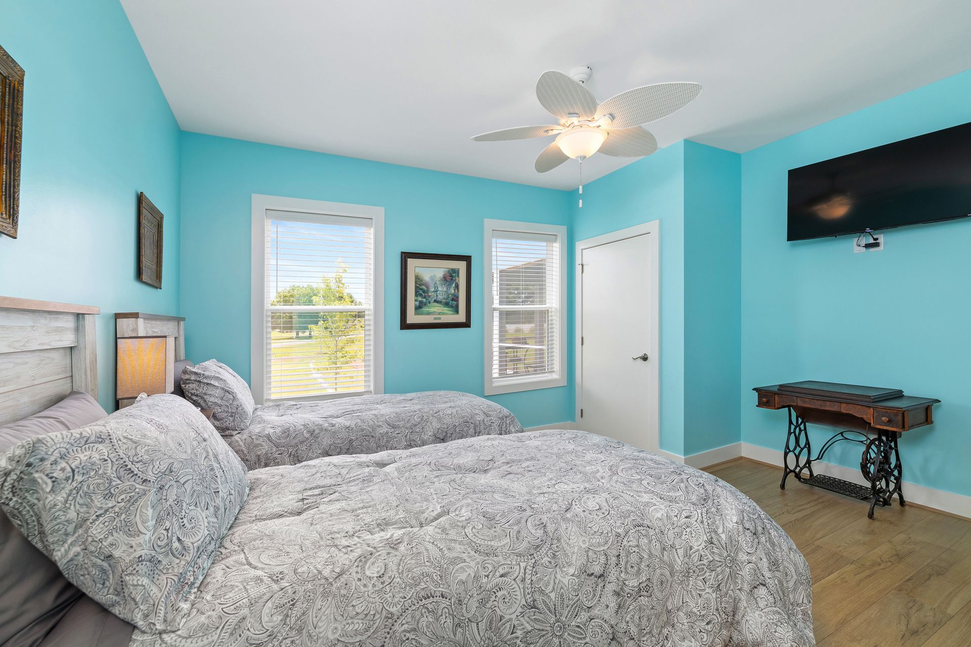 Two twin beds with fluffy gray blankets in a bedroom with light blue walls, two windows, and a mounted TV.