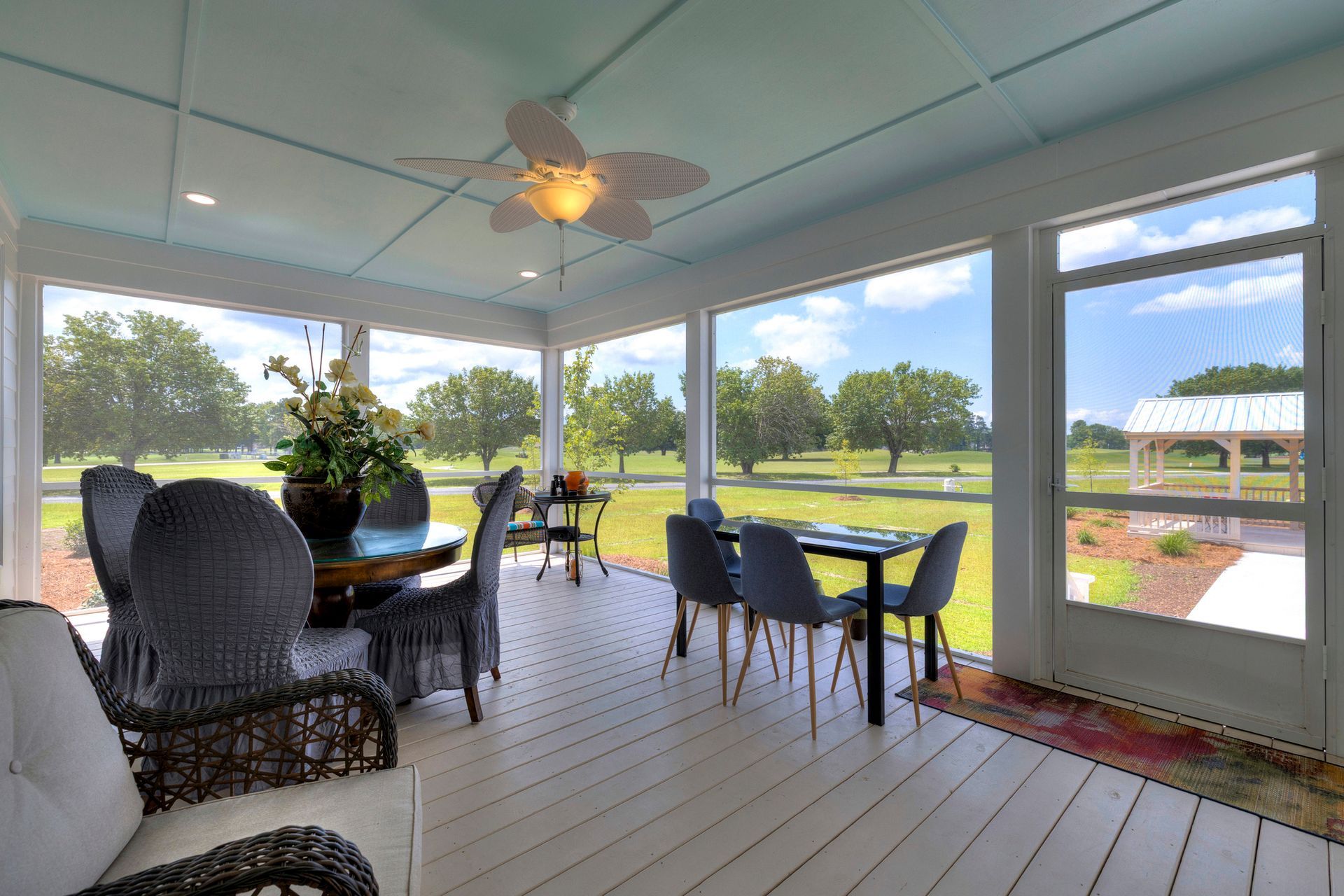 Screened porch with dining tables and chairs, offering views of a green field and trees under a blue sky.