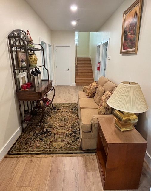Long hallway with light-colored walls and wood flooring. A sofa, desk, and rug with art and decorative items are along the sides. Stairs are visible in the distance.