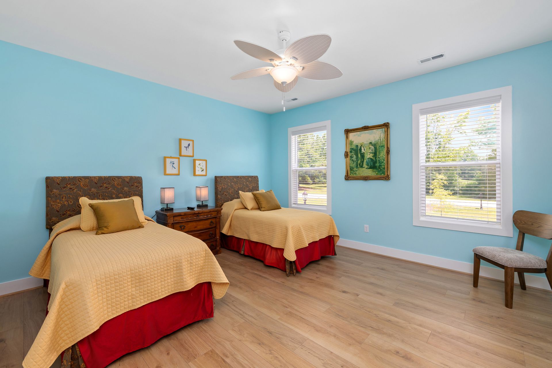 Bedroom with two twin beds, light blue walls, and a ceiling fan. Each bed has a yellow quilt and a red bed skirt.