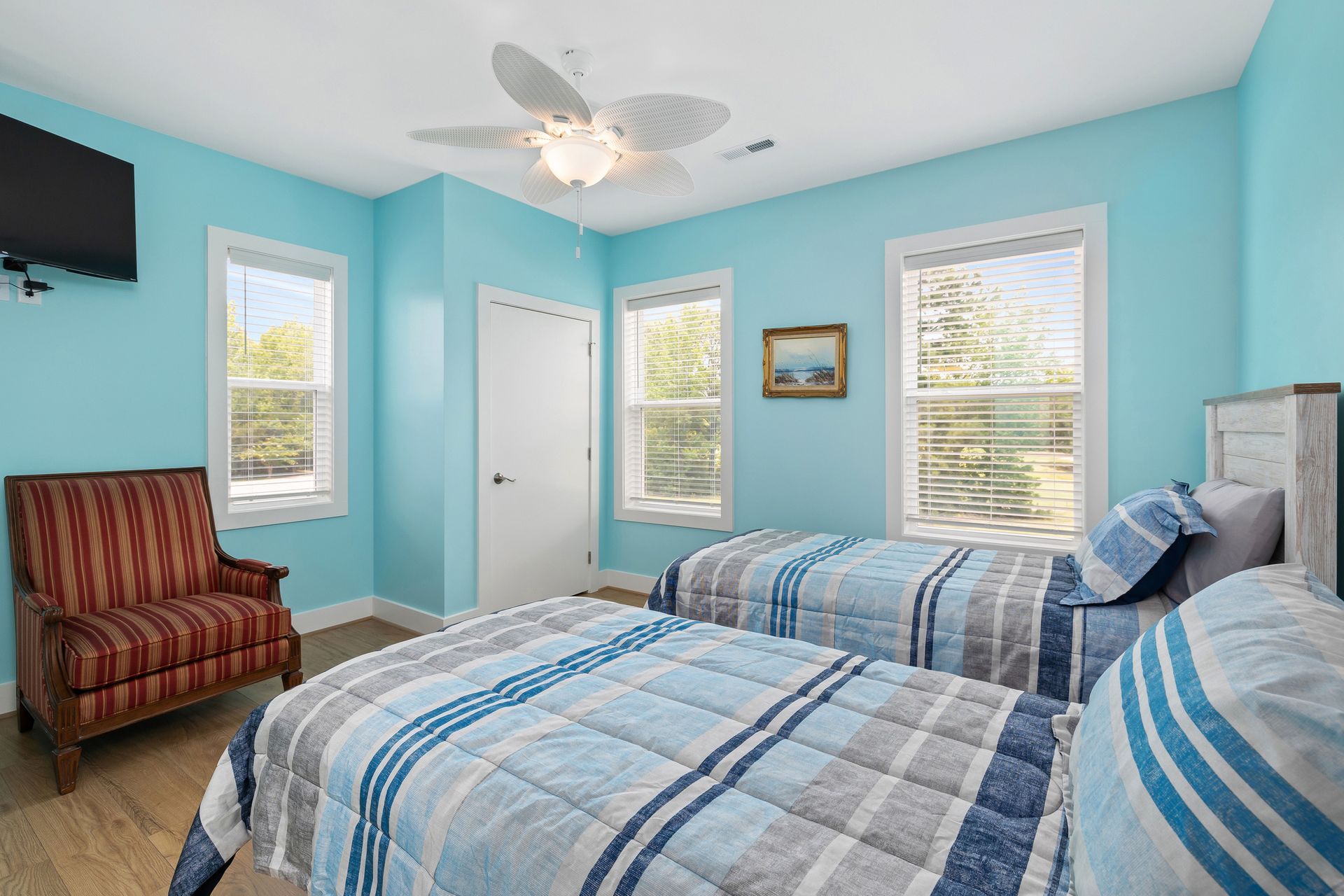 Bedroom with two twin beds, turquoise walls, a patterned armchair, and windows with sunlight.