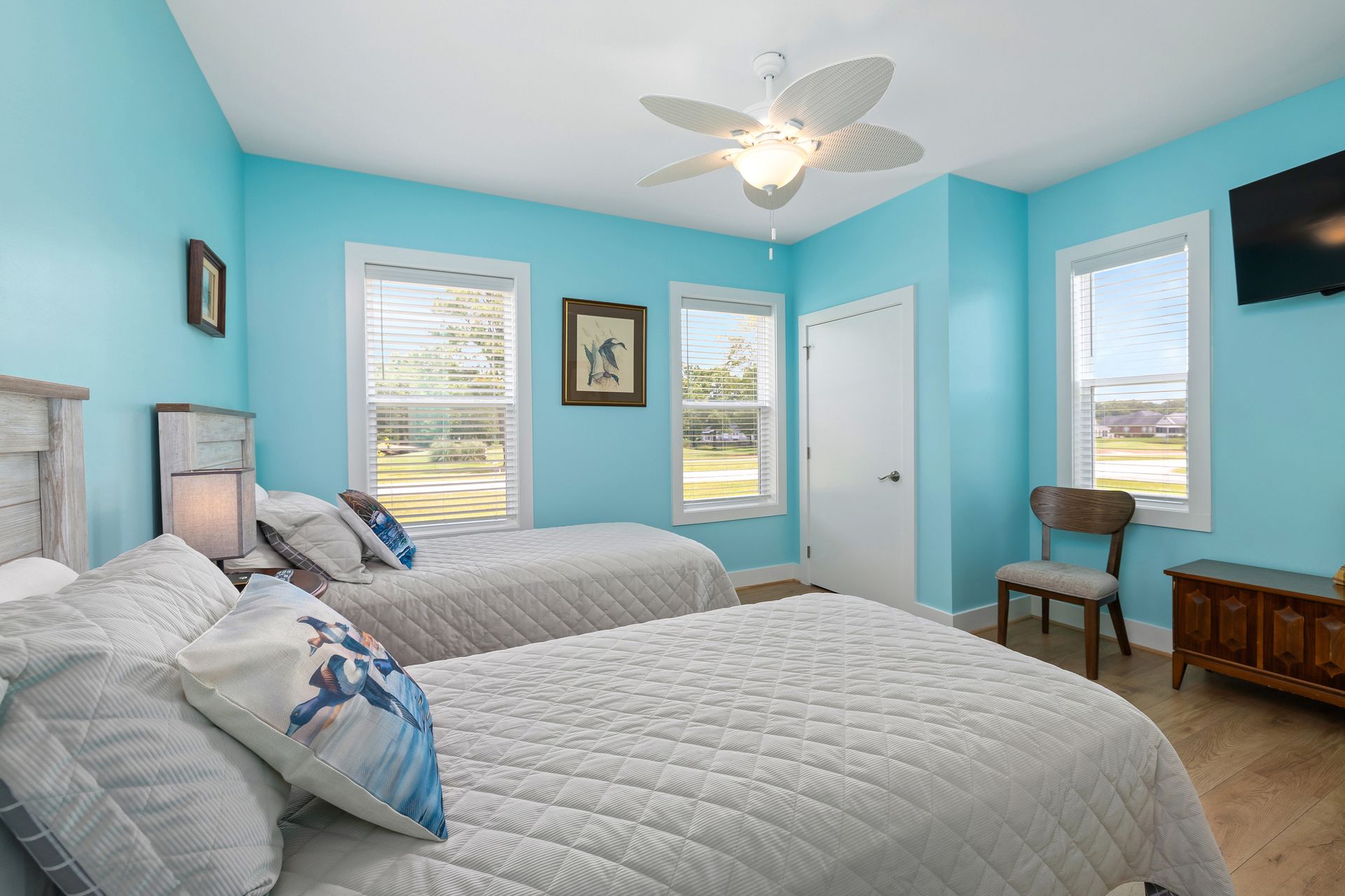 A bright bedroom with two twin beds, blue walls, and a ceiling fan. There are two windows, a chair, and a TV.