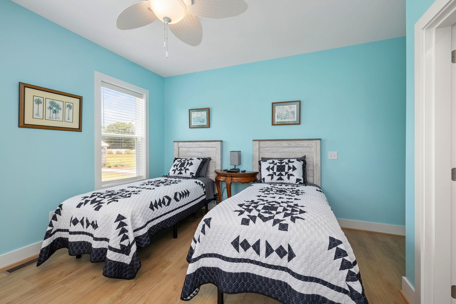 Two twin beds with white and navy quilts in a turquoise bedroom, with artwork and a ceiling fan.