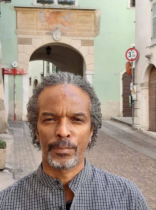 Man with graying hair and beard stands in front of an archway in a European street.
