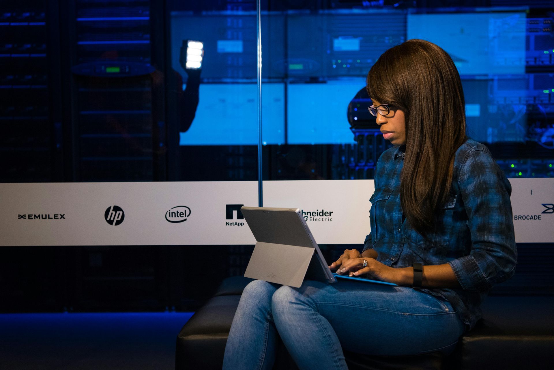 Woman with glasses and a blue plaid shirt sits on a bench, typing on a tablet in a data center.
