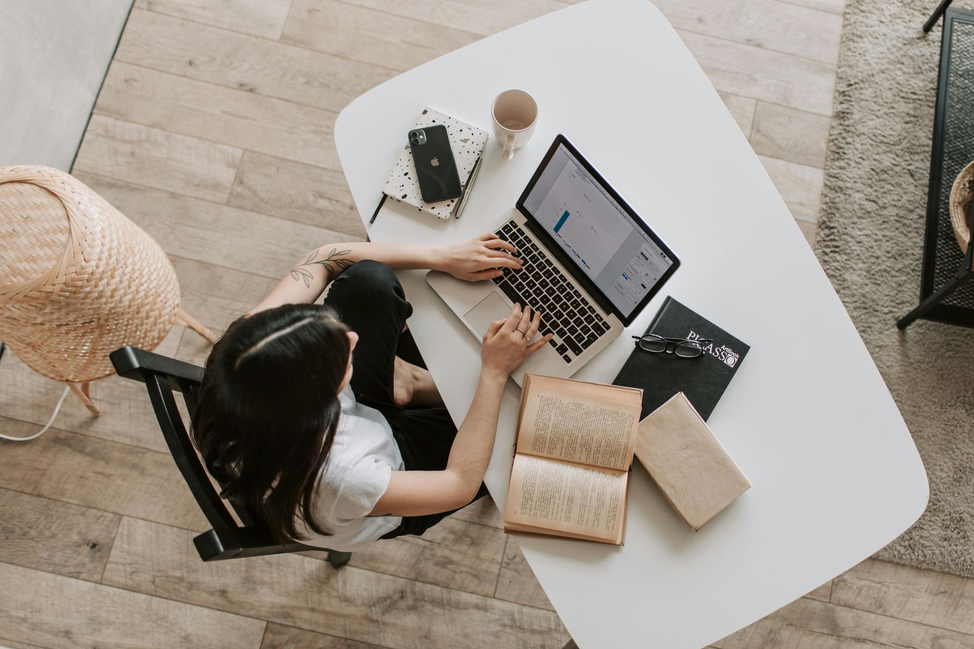 Woman working on a laptop at a white desk with books and a coffee cup. She is in a bright room with wooden floors.