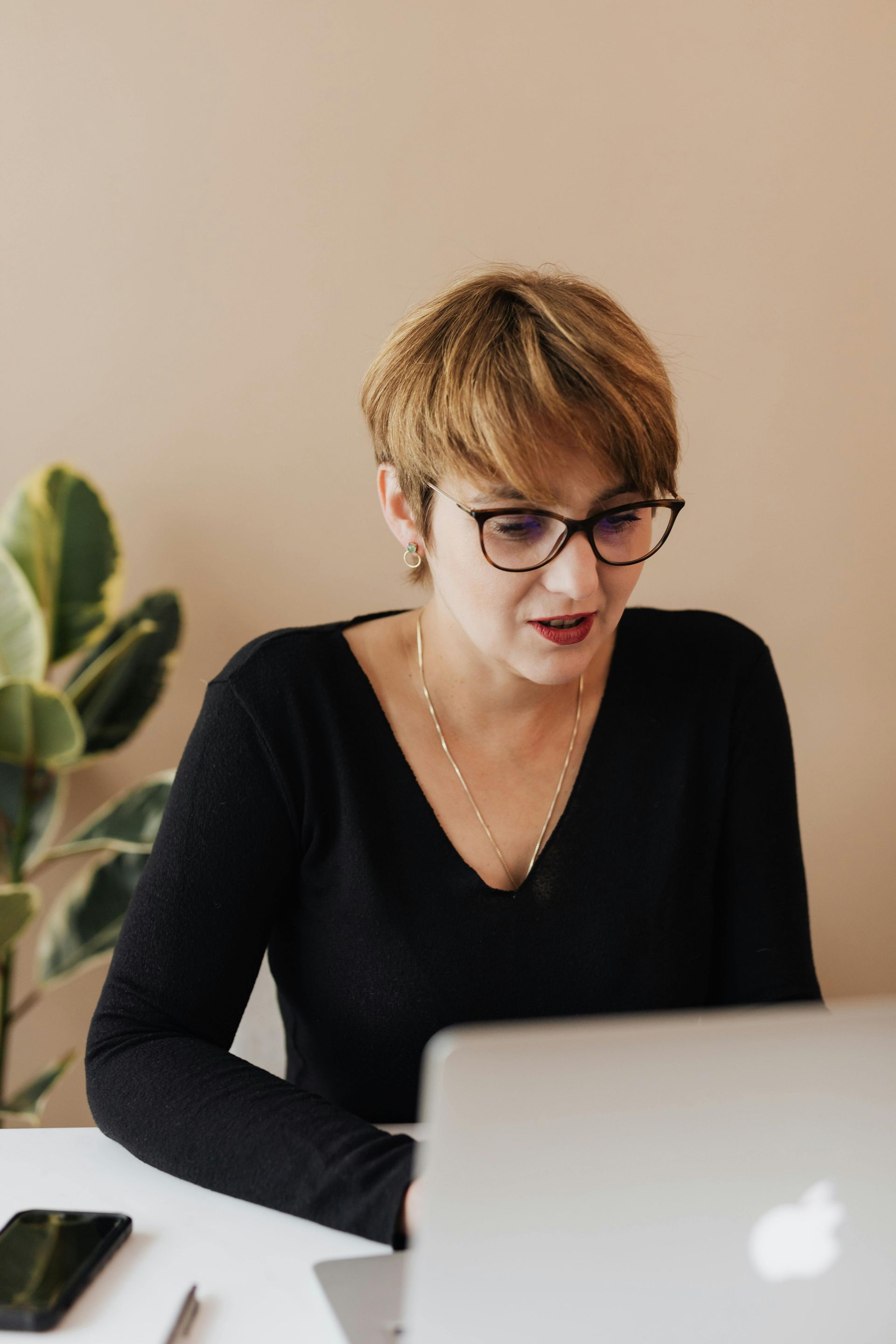 Woman with glasses working on a laptop at a desk, facing the screen. Plant and phone are nearby.