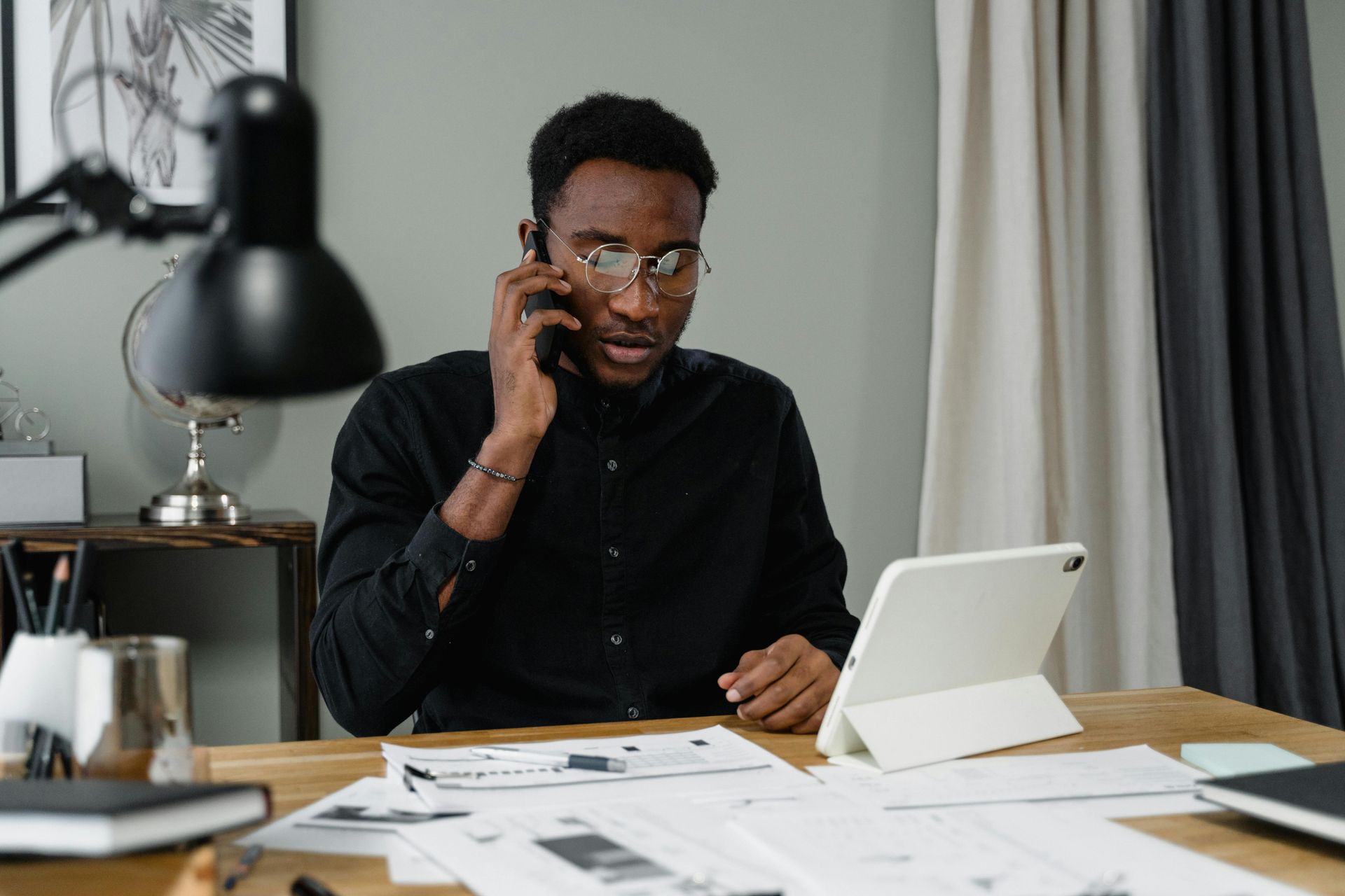 Man on phone at a desk with tablet, papers, and lamp. Black shirt, glasses, focused expression.