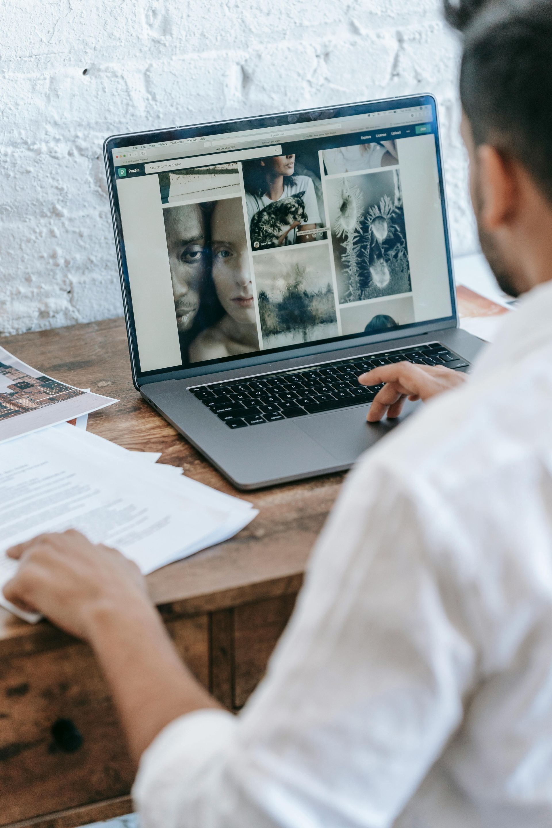 Man sitting at a desk, using a laptop displaying photos while looking at papers.