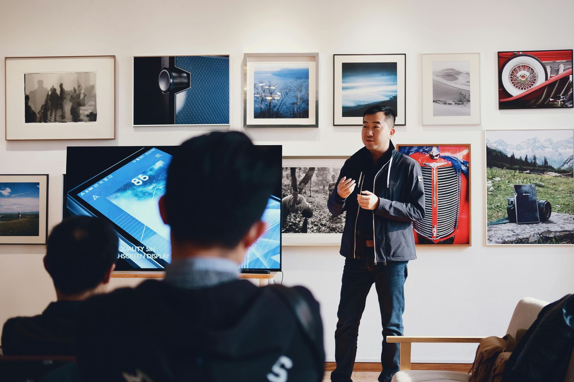 Man presenting, standing in front of a screen and audience. Photos hang on the wall.