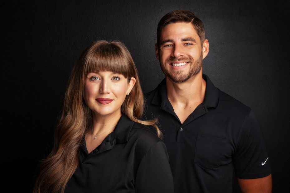 Woman and man in matching black shirts smile, dark background.