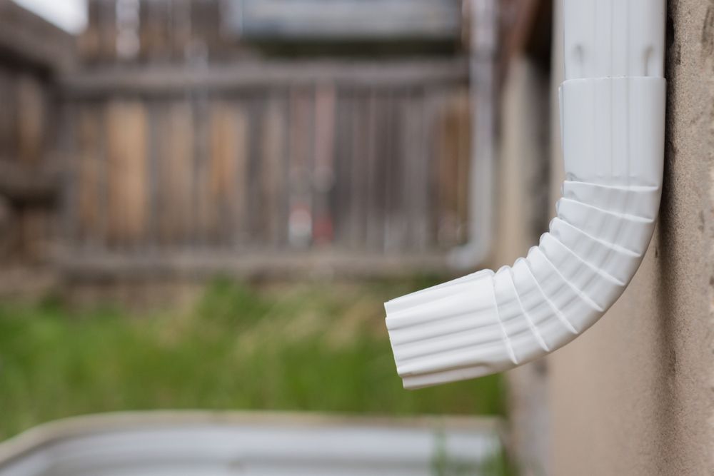 White rain gutter elbow on beige building exterior, blurred wooden fence and green grass in background.