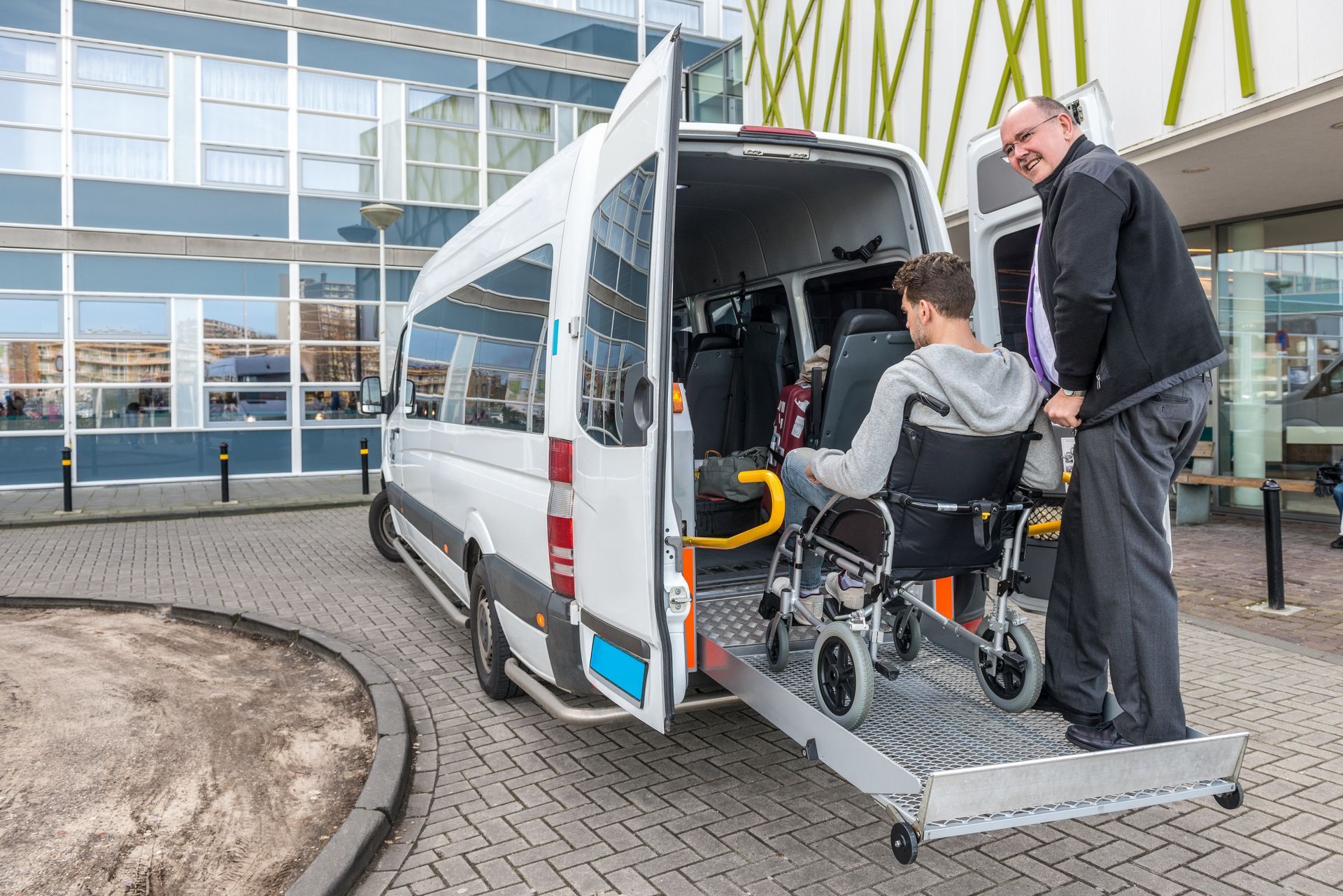 A driver assists a person in a wheelchair as they board a white accessible van using a hydraulic lift at a building entrance.