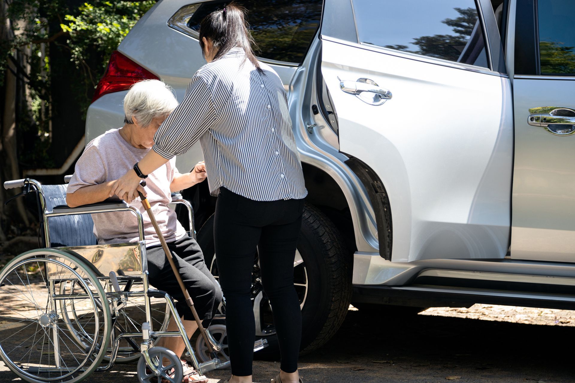 A person assisting an individual from a wheelchair into the passenger seat of a silver SUV parked outdoors.