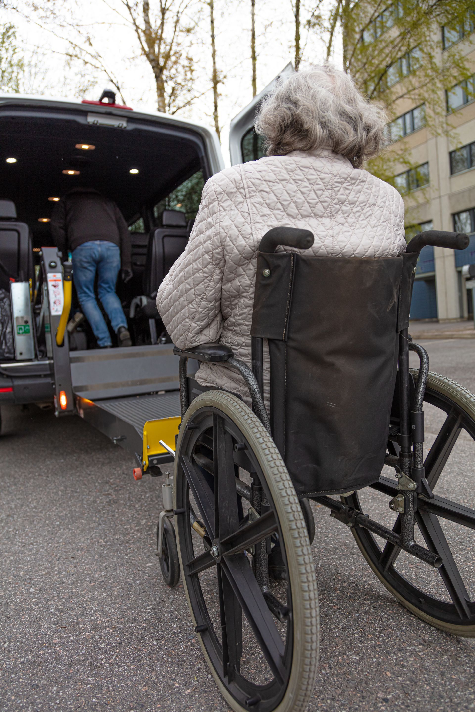 A person in a wheelchair waits as a driver enters an accessible van with a rear ramp deployed.
