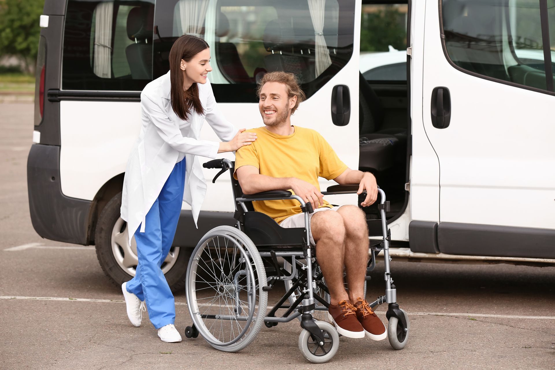 A smiling medical professional in scrubs placing a hand on the shoulder of a person seated in a wheelchair by a van.