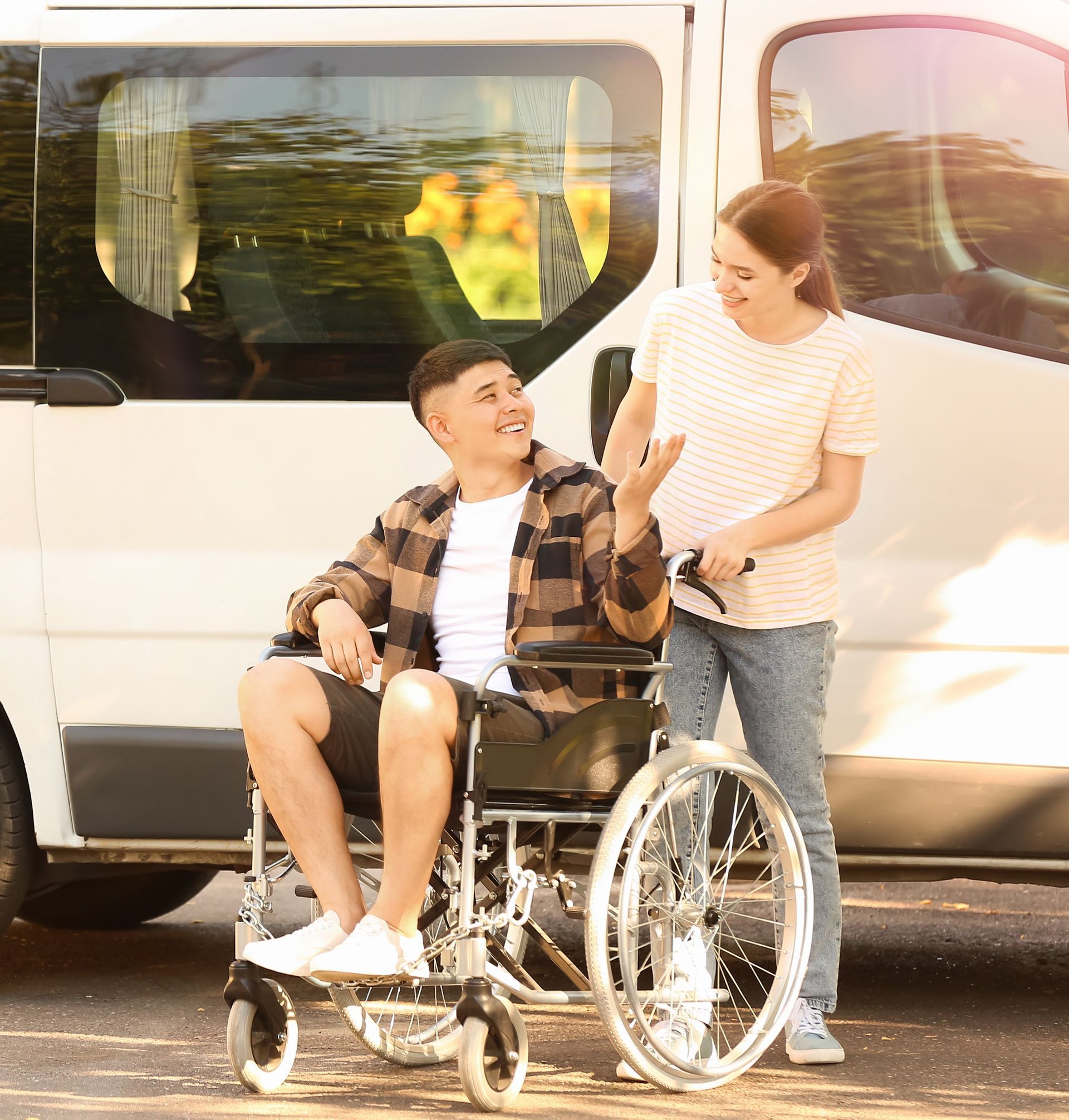 A person in a wheelchair smiles while talking to an attendant standing next to a white van in an outdoor setting.