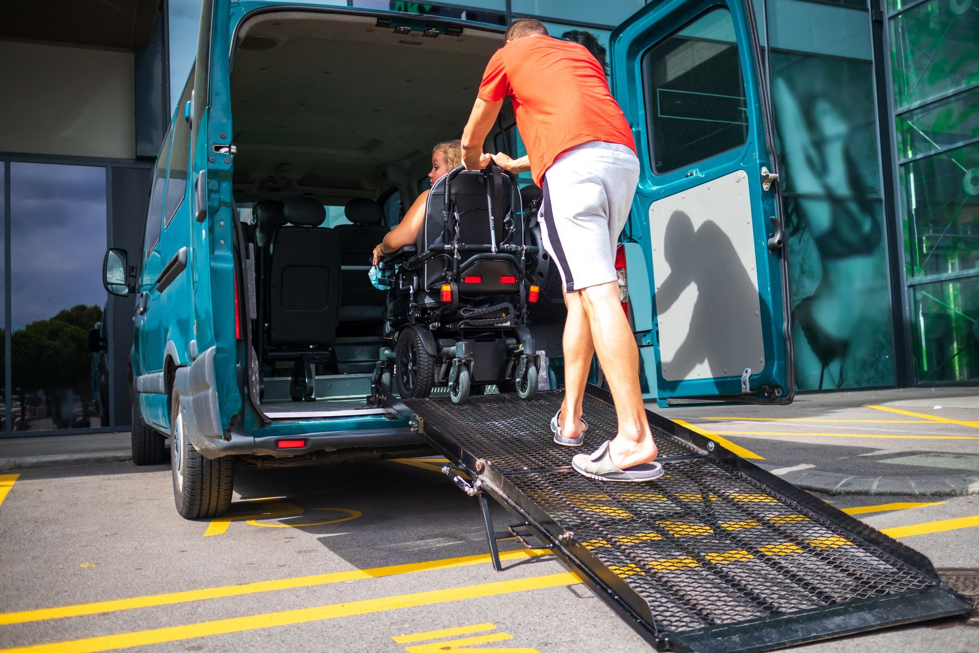 A person in a wheelchair is being assisted up a ramp into a teal van parked in an accessible parking space.