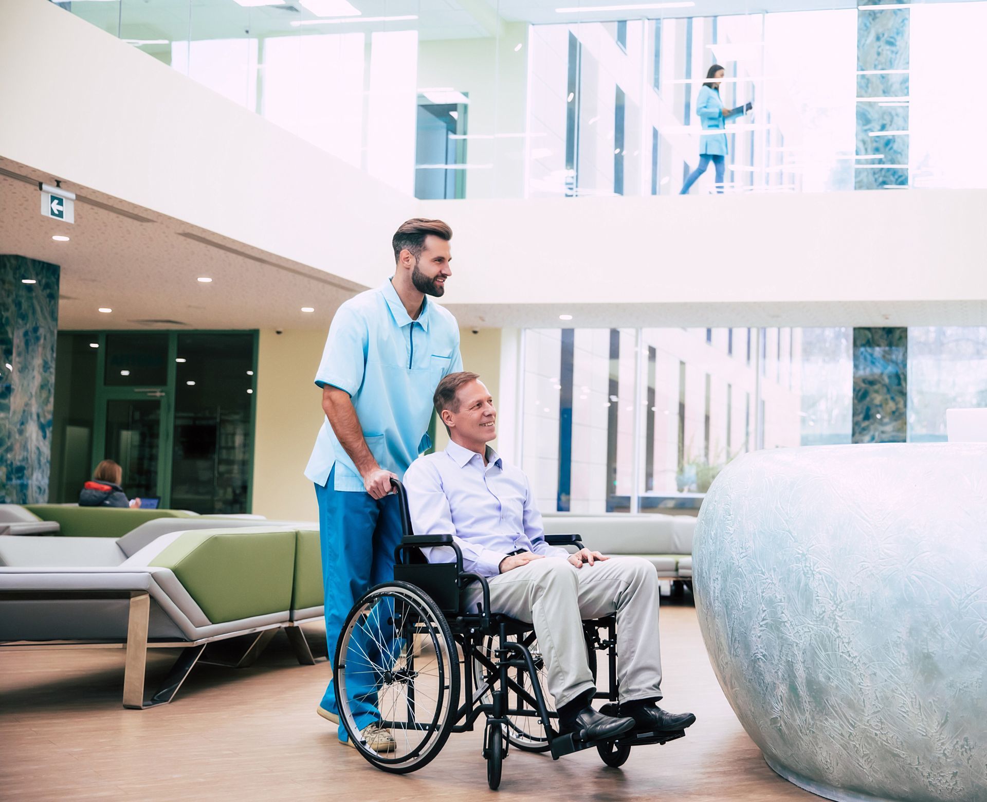 A caregiver in blue scrubs pushes a person in a wheelchair through a modern, brightly lit lobby.