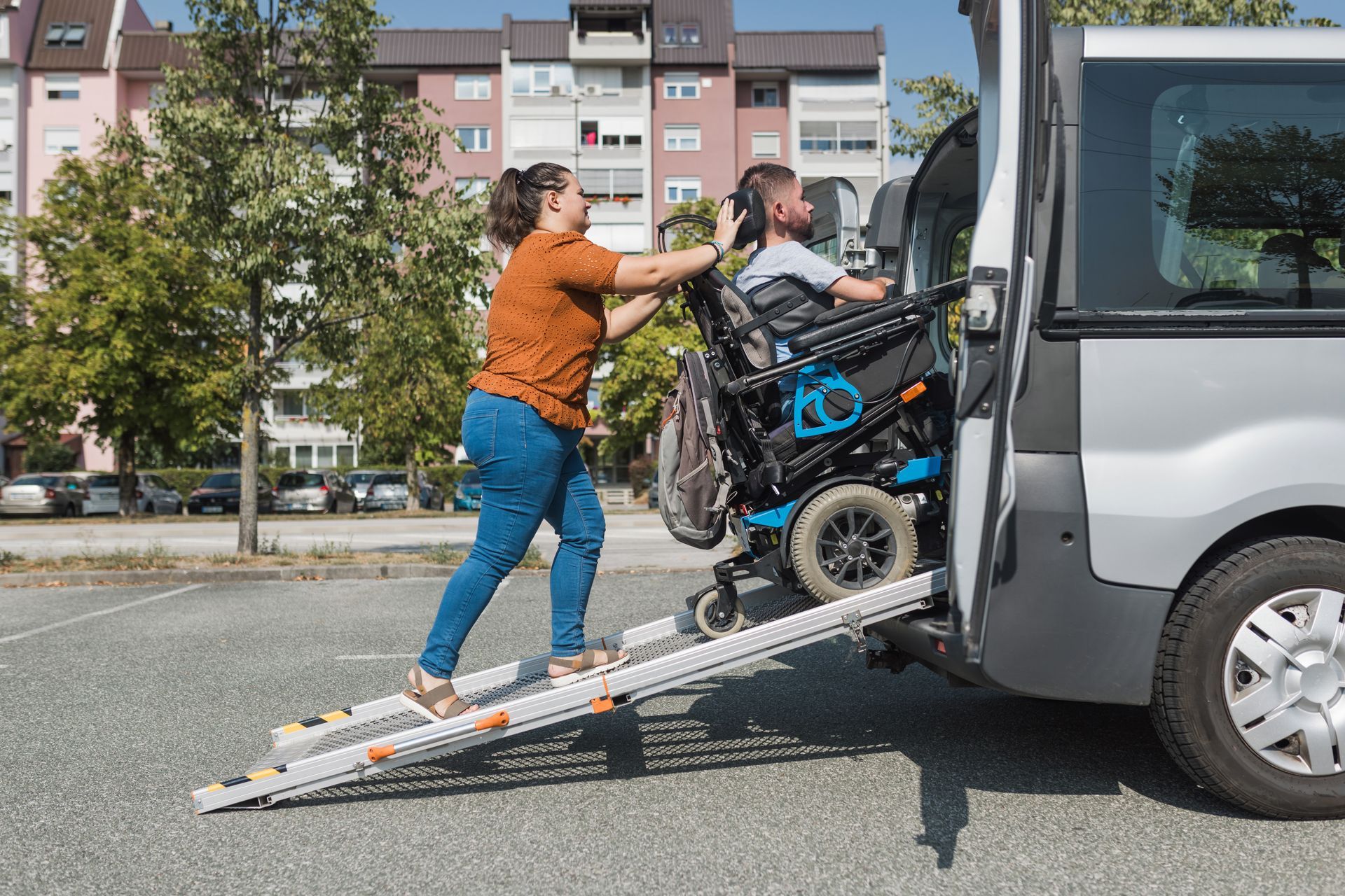 A person pushing a manual wheelchair up a portable ramp into the back of a van in a parking lot.