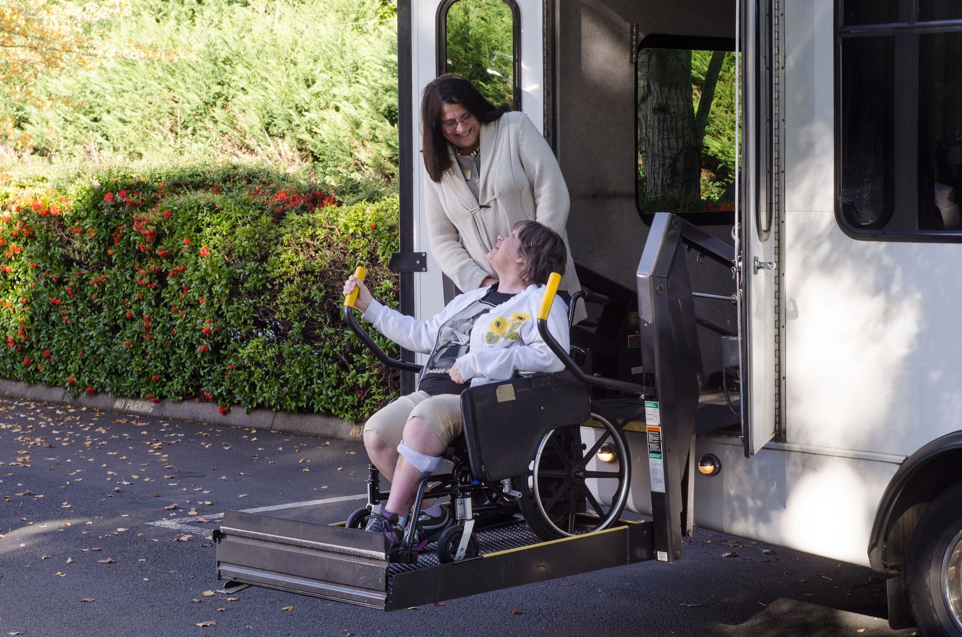 A person in a wheelchair uses an electric lift to board a shuttle bus, accompanied by someone standing in the doorway.