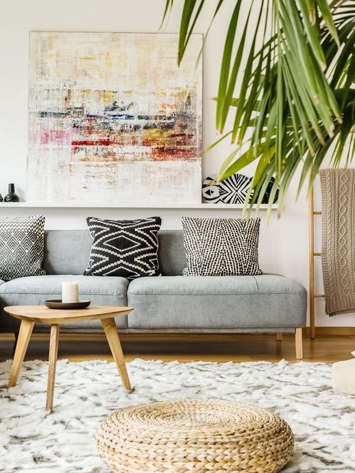 Living room with gray sofa, abstract art, textured rug, and decorative pillows. A wooden coffee table and large plant are also present.