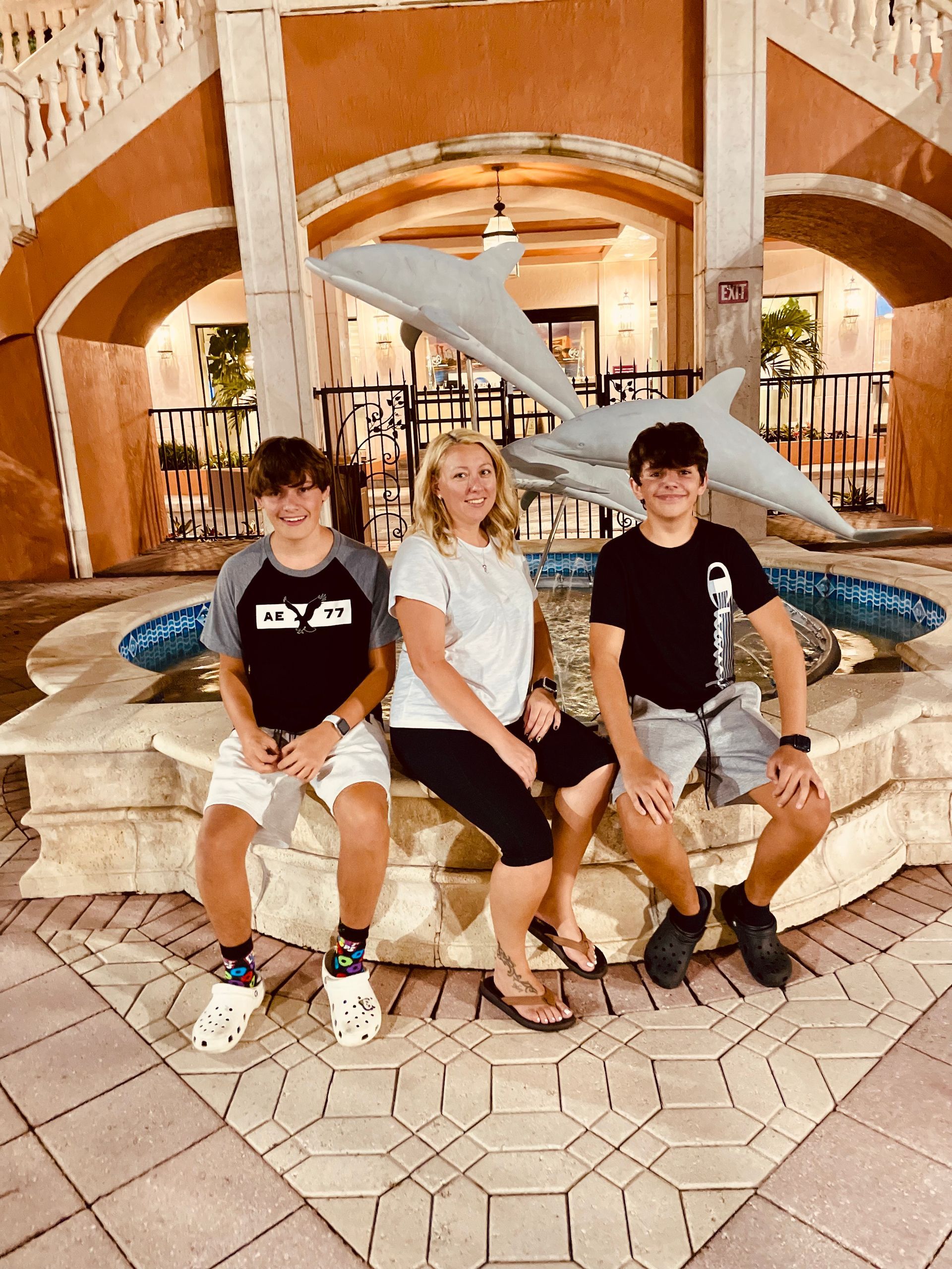 A woman and two young men sit on a stone fountain with a dolphin sculpture, outdoors at night.