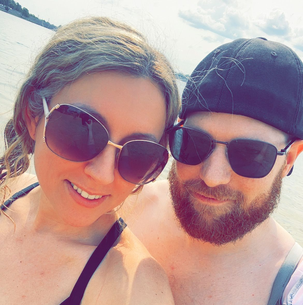 Couple in sunglasses smiling, near water on a sunny day. Woman's arm around man.